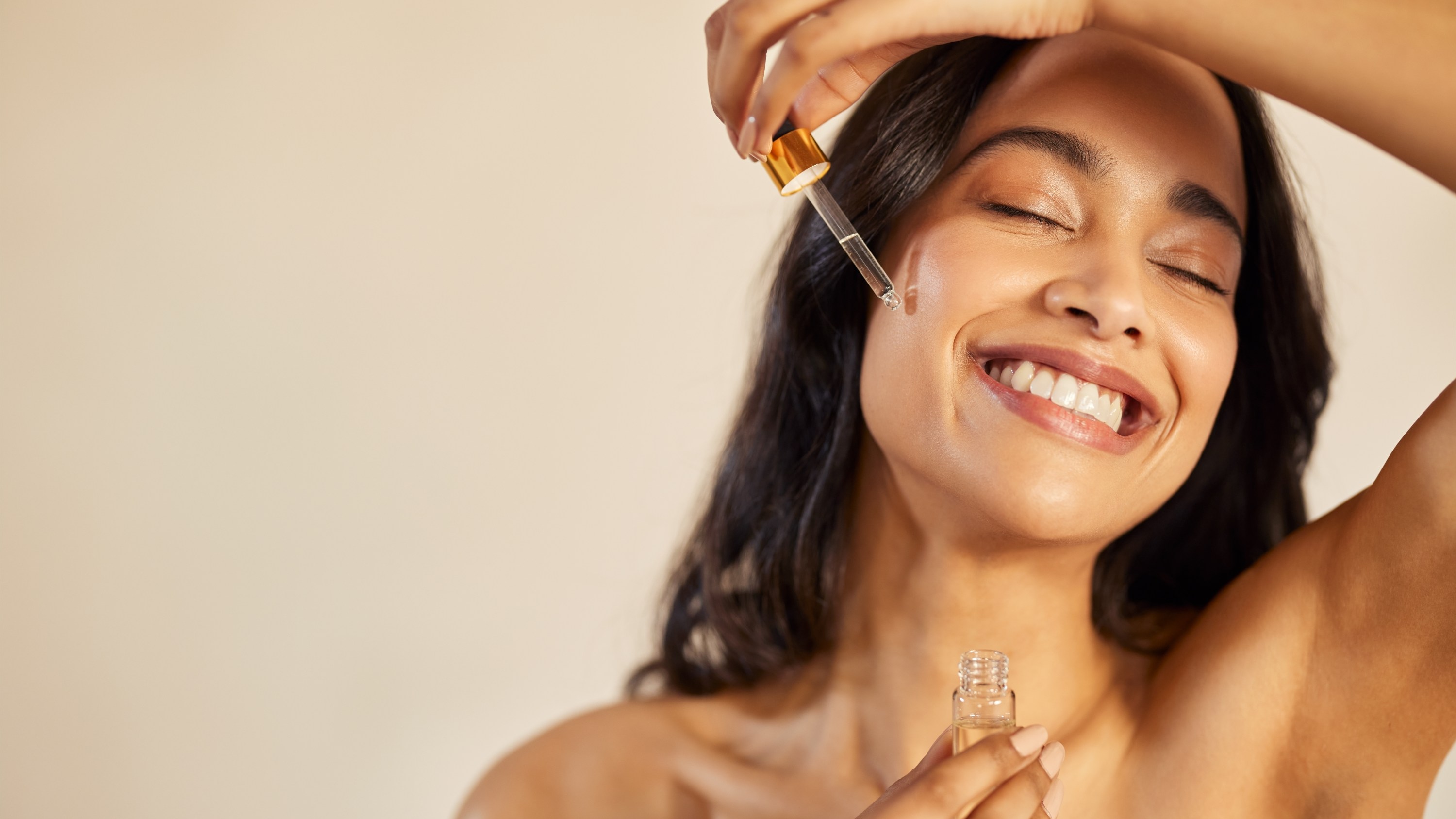 Happy young woman with bare shoulder applying serum on face with closed eyes. Beautiful hispanic young woman moisturizes her skin with serum isolated against beige background with copy space. Healthy and skin care conscious girl using facial treatment in summer.