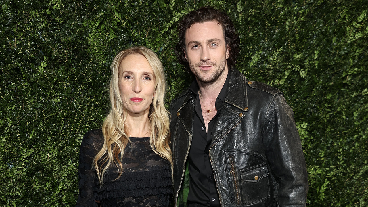 Aaron Taylor Johnson, a white man with brown hair and a beard, wearing a leather jacket posing with Sam Taylor-Johnon who is wearing a blazer, black top and bag.