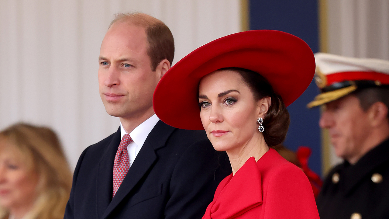 Prince William, a white man with blonde hair wearing a suit, and Kate Middleton, a white woman with a red dress and hat, staring in a royal manner.