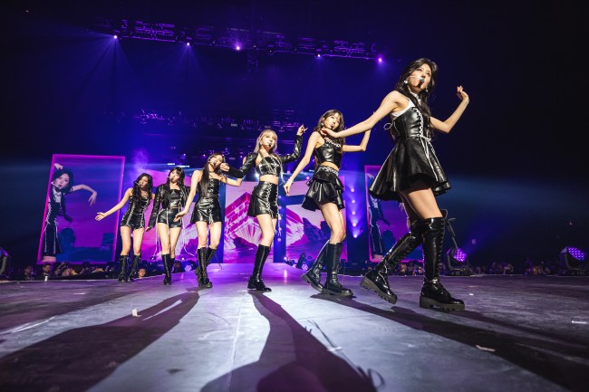 K-Pop Girl Group IVE, six Asian women ANYUJIN, GAEUL, REI, JANGWONYOUNG, LIZ, and LEESEO, dancing with black leather outfits on stage. 