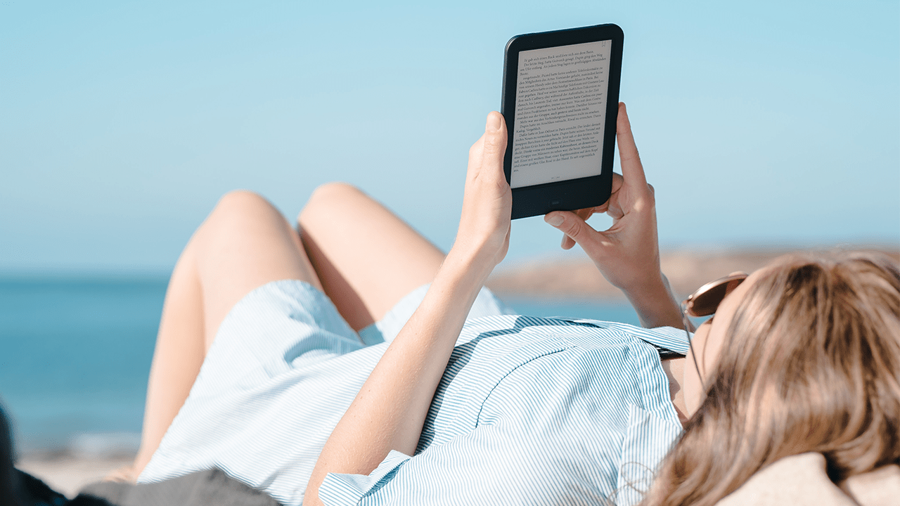 Woman laying down and reading on her Amazon Kindle device on the beach.