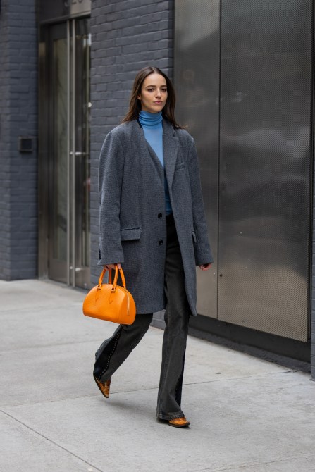 A woman wears a gray wool coat, orange bag, blue turtleneck, and gray denim on February 10, 2024 in New York City.