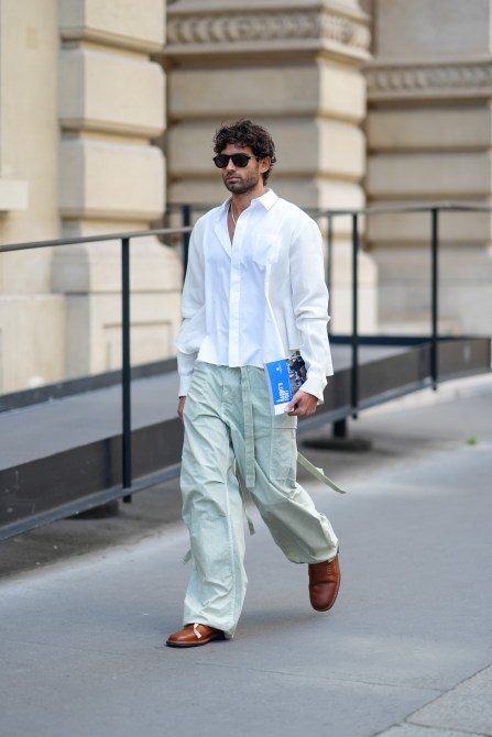 A man wears black sunglasses, white necklace, a white button long sleeve shirt, light green cargo loose pants, shiny light brown boots leather shoes