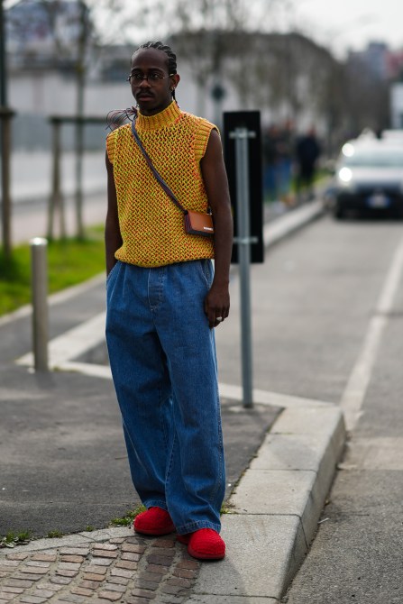 A guest wears glasses, an orange / yellow / red braided wool sleeveless pullover, a camel shiny leather accordion crossbody bag from Sunnei, blue denim large jeans pants, red embossed pattern shoes, outside the Sunnei fashion show, during the Milan Fashion Week Fall/Winter 2022/2023 on February 24, 2022 in Milan, Italy.