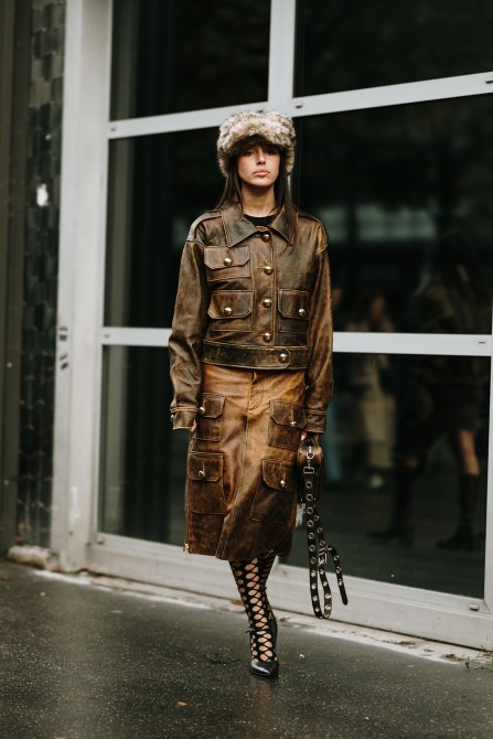 a woman wears a brown grungy leather jacket and matching skirt with a furry hat and lace up heels.