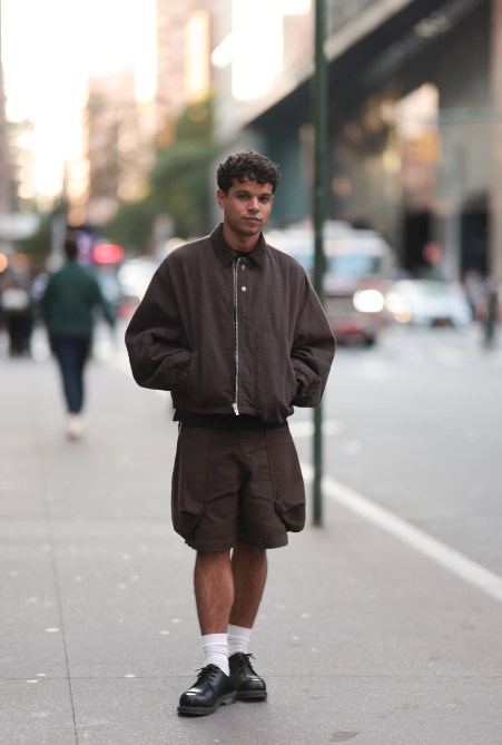A Fashion Week guest is seen wearing two tiny stud earrings; a dark brown blouson jacket with a corduroy collar, puffy sleeves, a silver zipper, and two silver buttons from Jacquemus; a black T-shirt underneath; knee-length dark brown cargo shorts with large pockets along the sides from Jacquemus; white socks; and black leather three-eye shoes with an exposed silver steel toe cap from Dr. Martens before the Jacquemus Boutique Store Opening during the New York Fashion Week on September 04, 2024 in New York City.