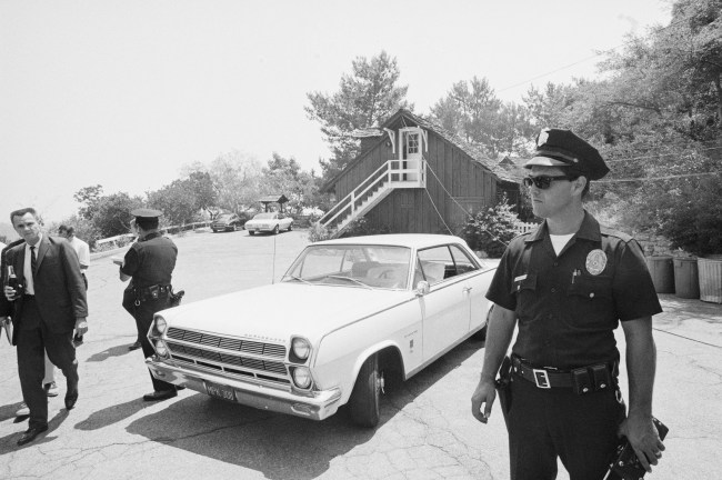 (Original Caption) Beverly Glen, California: Police officer stands in front of a car containing the sheet covered body of one of the five persons found dead in and around the barn type home of actress Sharon Tate. Tentatively identified as victims are: actress Sharon Tate and hairdresser Jay Sebring, found inside the house, photographer Voy Frykoski and coffee heiress Abigail Folger -- found on the lawn under trees at the far left -- and the man in the car who has not been named.