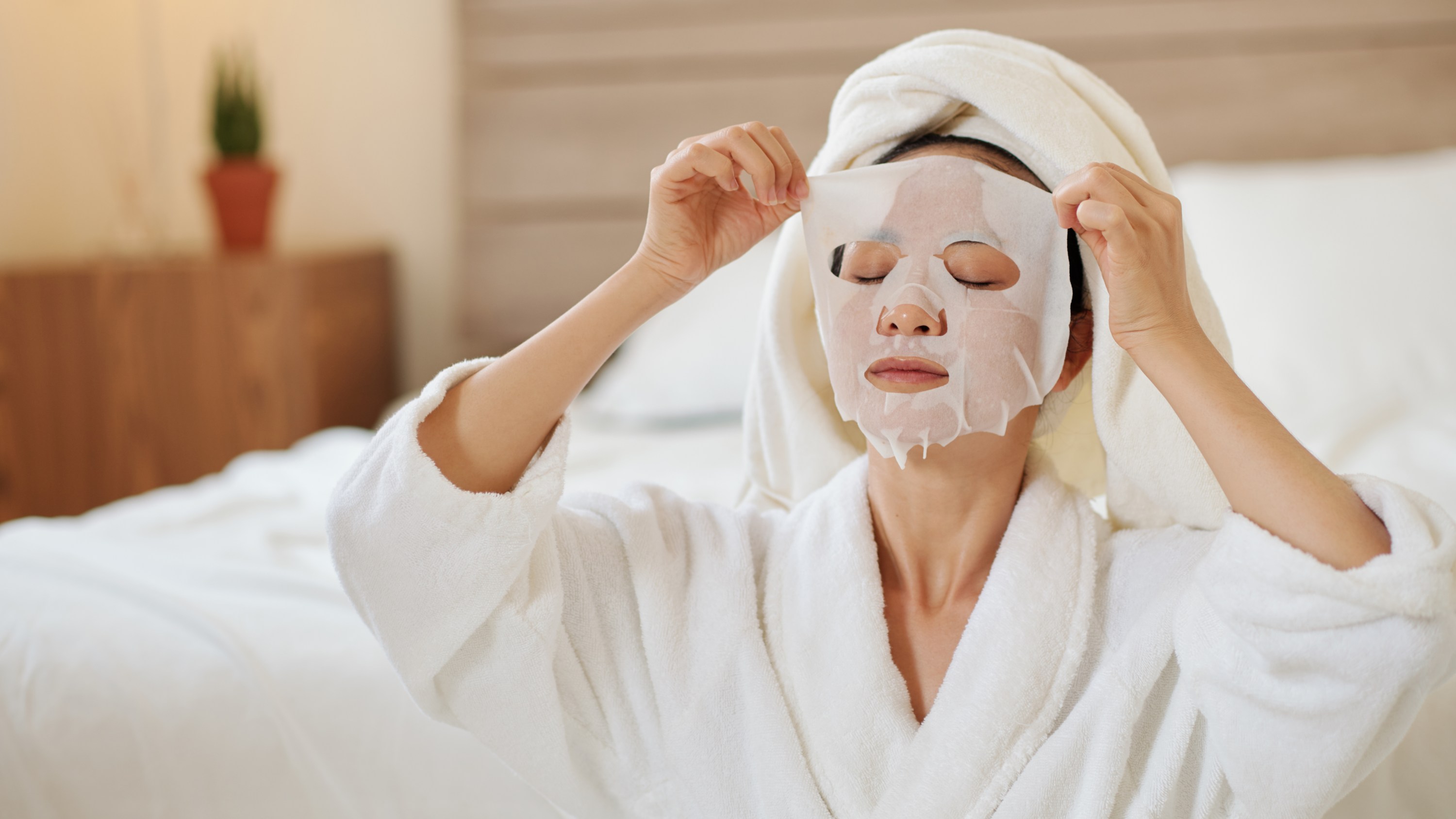 Young woman in bathrobe taking off moisturizing sheet mask