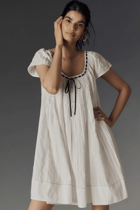 A model wearing the By Anthropologie Tie Mini Swing Dress in vanilla