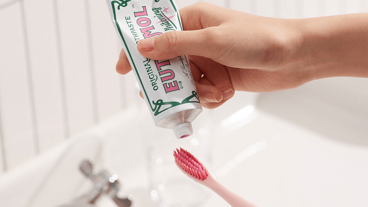 A hand squeezing Euthymol's Pink Whitening Toothpaste onto a pink toothbrush over a white sink
